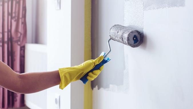 Hand in a yellow protective glove painting a wall in apartment in gray colour with a roller brush.