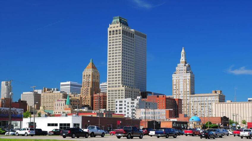 "A view of Tulsa downtown buildings, including three historic buildings and two skyscrapers, with a parking lot and cars in the foreground.