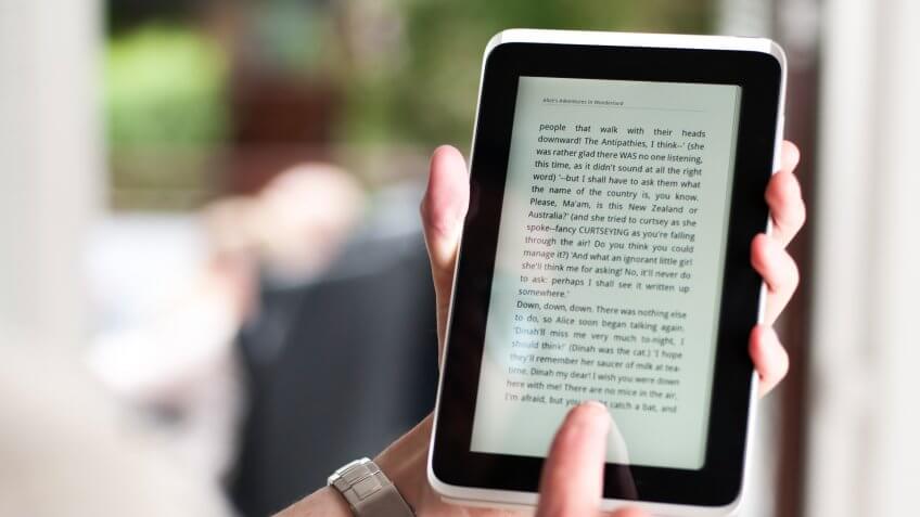 Left hand is holding a tablet or ereader with a digital version of the novel Alice's Adventures In Wonderland by Lewis Carroll.