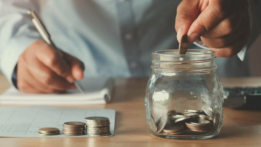 man putting coins in a jar while writing in a notebook