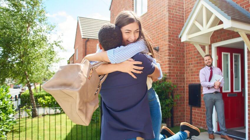 a couple embrace in front of their new home , in the background an estate agent looks on happily.