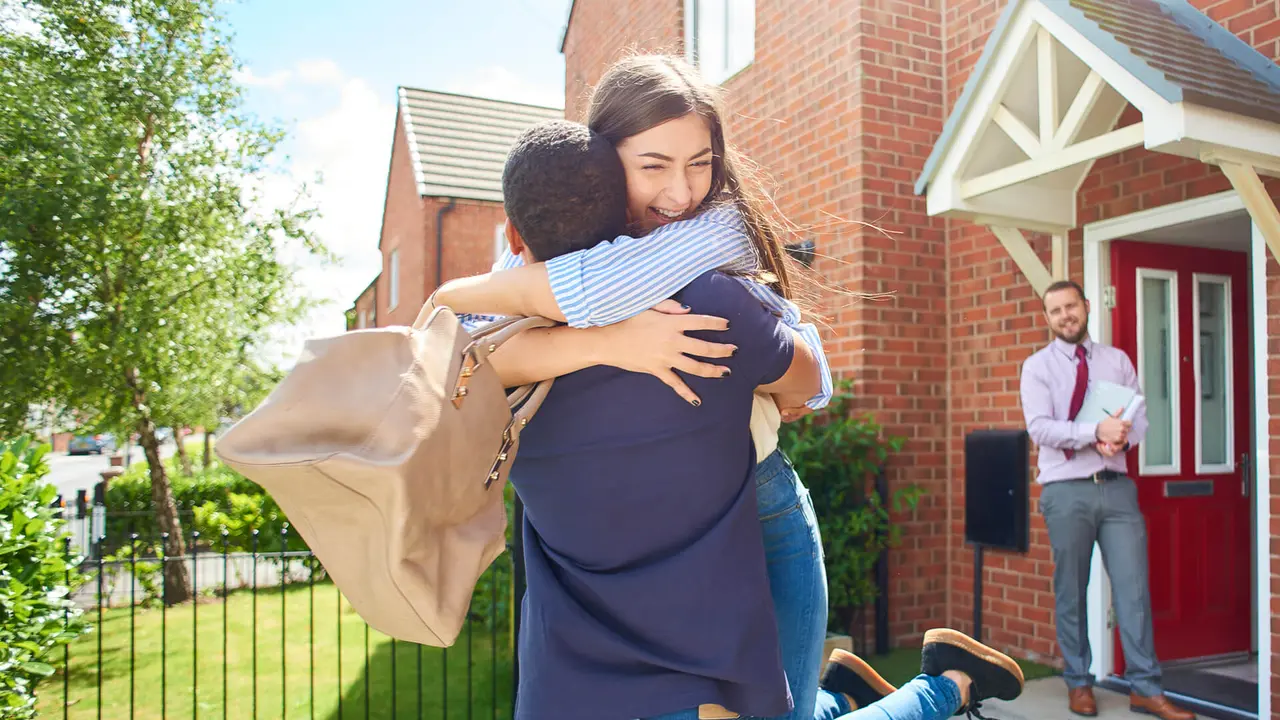a couple embrace  in front of their new home , in the background an estate agent looks on happily.