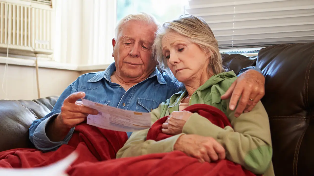 Worried Senior Couple Sitting On Sofa Looking At Bills Under A Blanket.