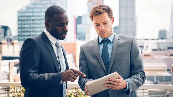 Afro american businessman discussing project on digital tablet with his business partner, standing outdoor, city scape in the background.