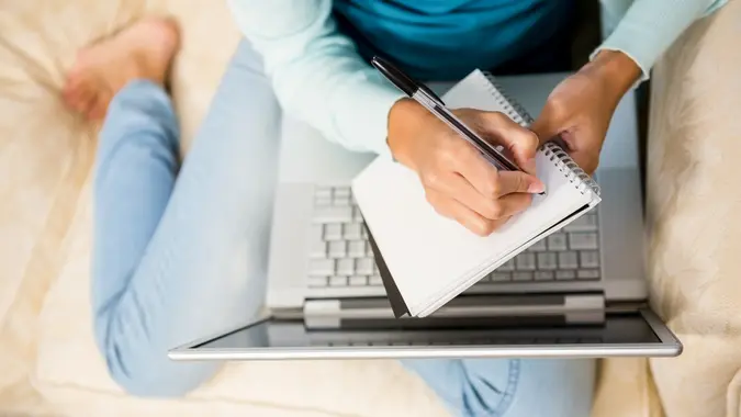 woman on a sofa writing in a notebook over a laptop