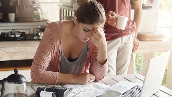 Thoughtful stressed young female sitting at kitchen table with papers and laptop computer trying to work through pile of bills, frustrated by amount of domestic expenses while doing family budget.