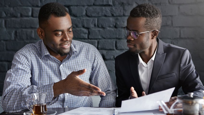 Businessman in glasses and suit holding papers, showing presentation of his project to cheerful business partner, who smiling happily, supporting his idea with positive gesture.