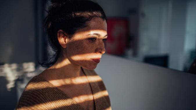 Portrait of a sad young woman in her apartment, with a sun on her face, that is breaking through the window blinds // wide photo dimensions.