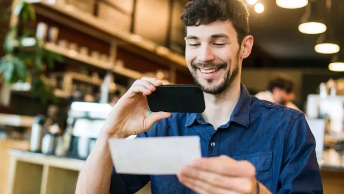 A smiling man takes a picture with his smart phone of a check or paycheck for digital electronic depositing, also known as "Remote Deposit Capture".