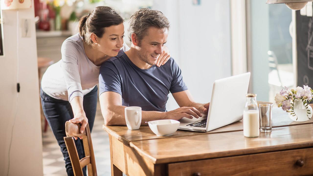 Nice thirty year couple using a laptop while having breakfast in the kitchen, they are wearing casual clothes and th man has gray hair.