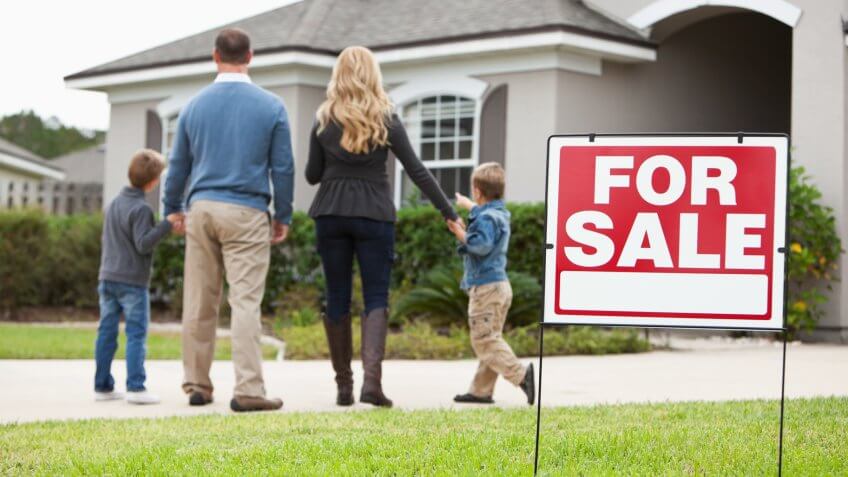 Family with two boys (4 and 6 years) standing in front of house with FOR SALE sign in front yard.