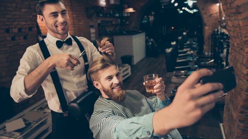 Smile! Cheerful handsome stylish red bearded guy is taking selfie photo at barber shop, classy dressed smiling stylist is making him a brand new haircut and posing also.