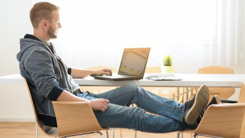 Young man working casual at his laptop with legs on the chair.