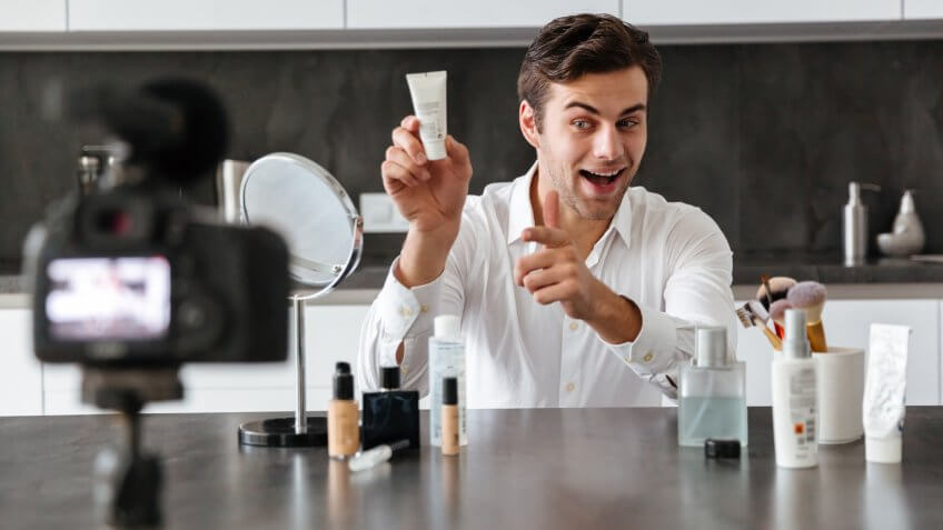 Handsome young man filming his video blog episode about new cosmetic products while sitting at the kitchen table and pointing finger.