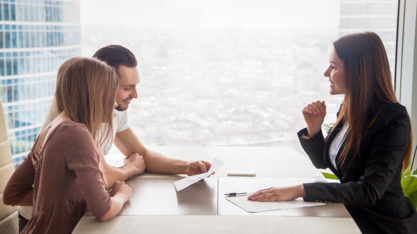 Side view of realtor and young couple sitting at office desk discussing property for sale.