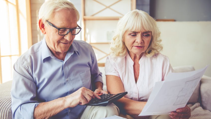 Handsome old man and beautiful woman are examining documents, using calculator and smiling while sitting on couch at home.
