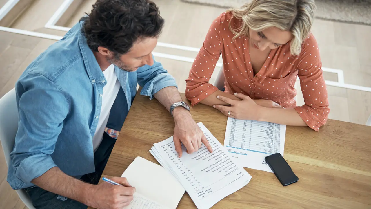 A couple sits at their kitchen table, which is covered in documents, as they review their financial paperwork together.