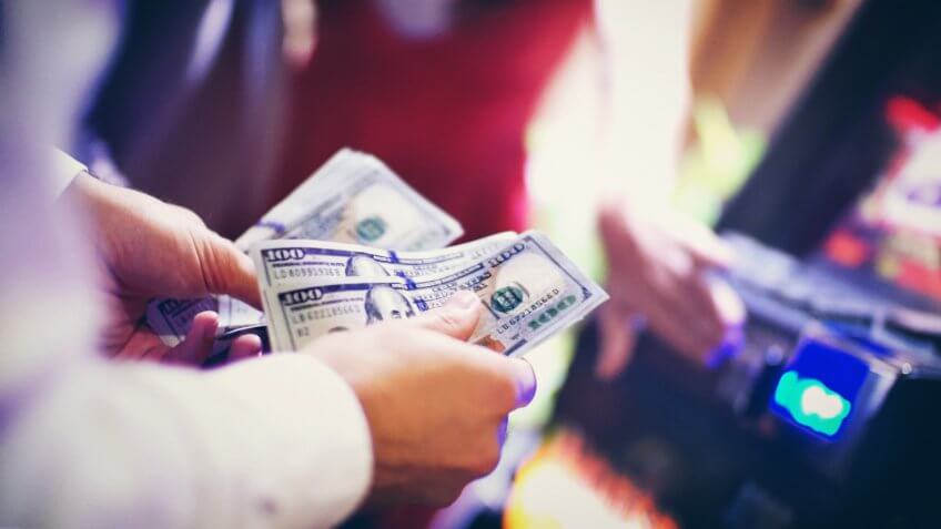 Closeup of unrecognizable man counting $100 bills in casino while some of his friends are still playing slots in background, blurry.