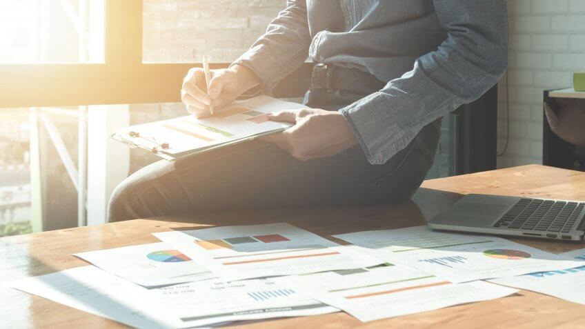 Businessman analyzing investment charts on document paper and using laptop computer at office.
