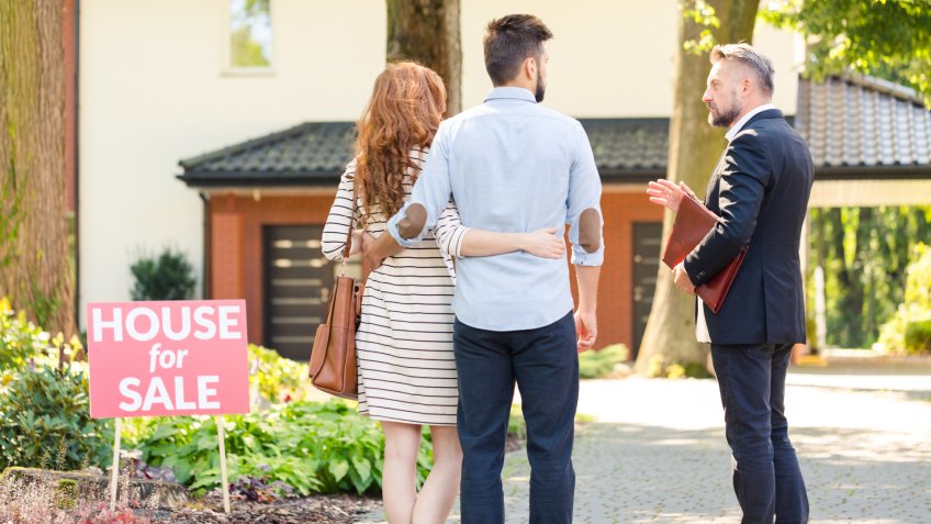 Real estate broker and clients standing next to plate with inscription "house for sale".