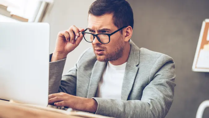 Handsome young man sitting at his workplace looking at his laptop and adjusting his glasses.