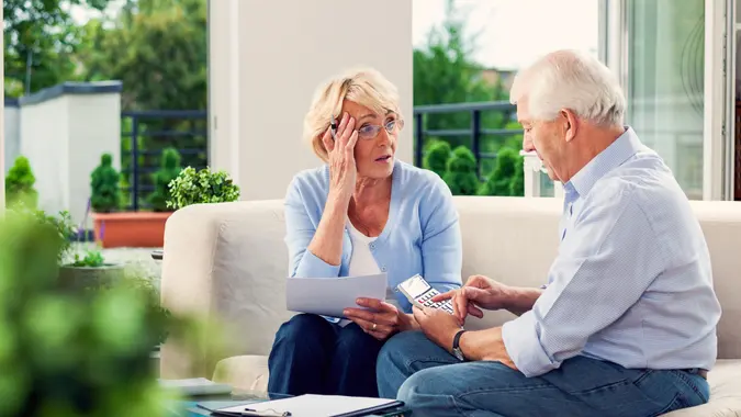 Worried senior couple sitting on sofa at home and looking over bills.