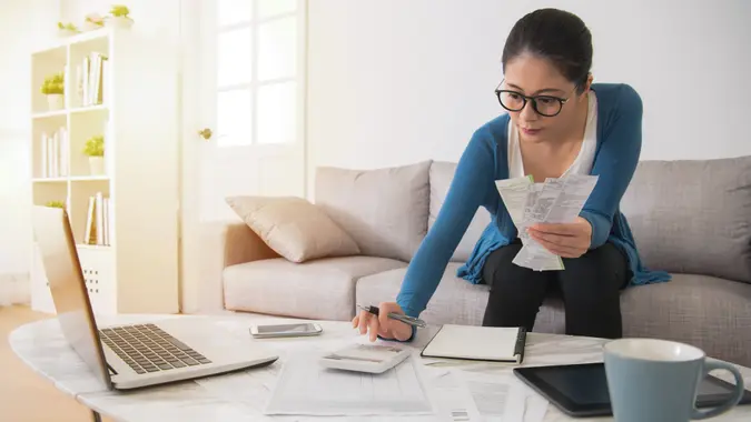 asian housewife calculating accounting new year budget holding a receipt using calculator sitting on sofa in the living room at home