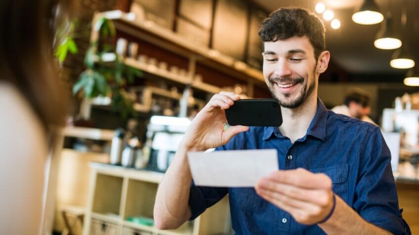 A smiling man takes a picture with his smart phone of a check or paycheck for digital electronic depositing, also known as "Remote Deposit Capture".