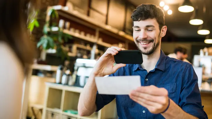 A smiling man takes a picture with his smart phone of a check or paycheck for digital electronic depositing, also known as "Remote Deposit Capture".