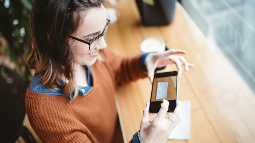 A smiling young woman takes a picture with her smart phone of a check or paycheck for digital electronic depositing, also known as "Remote Deposit Capture".