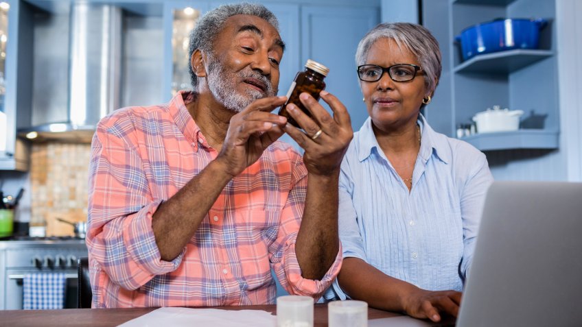 Man looking at medicine while sitting by woman using laptop in kitchen at home.
