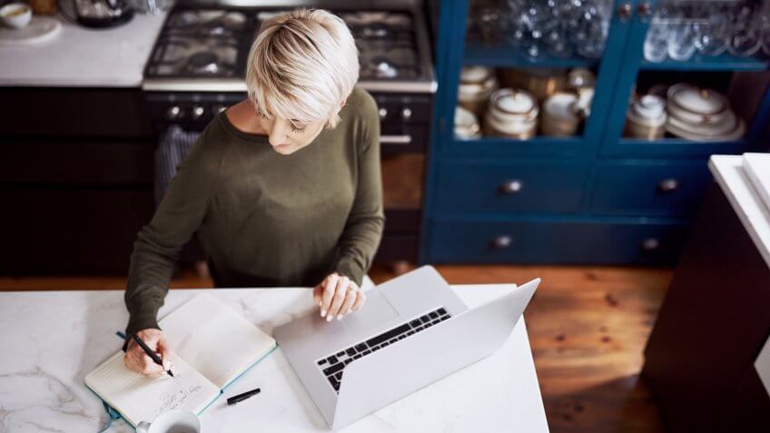 High angle shot of a mature woman writing in a notebook while working on a laptop at home.