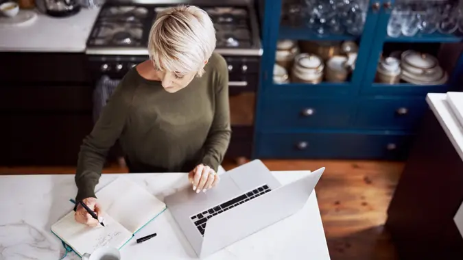 High angle shot of a mature woman writing in a notebook while working on a laptop at home.