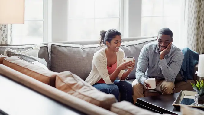 A young man and woman sit on a sofa, drinking coffee. They are smiling and relaxing in their living room.