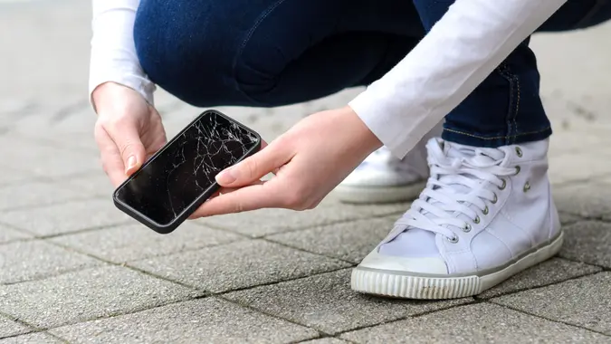 Close up view on kneeling person in jeans and shoes picking up broken phone on stone paved sidewalk outdoors.