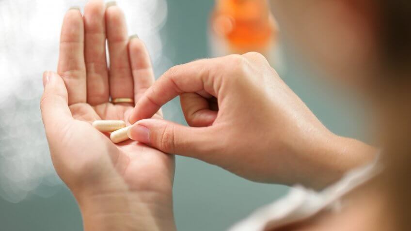 Close up view of young woman holding ginseng vitamins and minerals pills in hand with capsule bottle on table.
