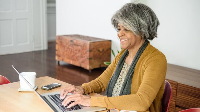 She is surfing the web at home, sitting at table typing on laptop keyboard.
