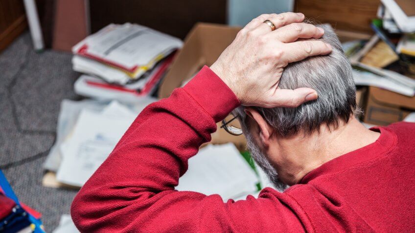 A senior adult man with attention deficit disorder (ADD) is completely frustrated and holding his head as he looks down at chaotic piles of notebooks and documents and paperwork scattered on the floor in his messy office space.