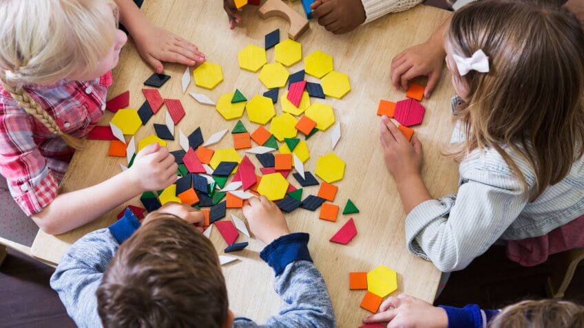 An overhead view of a group of five preschoolers sitting at a table playing with colorful blocks and geometric shapes.