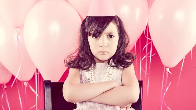 A young girl dressed for a party, standing in front of pink balloons with her arms crossed and a serious expression.