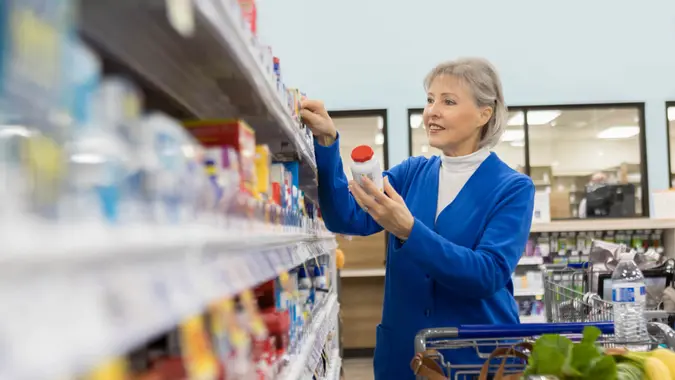 A senior woman stands in the aisle of a pharmacy and reads the pricing of an over the counter medication on the shelf label.