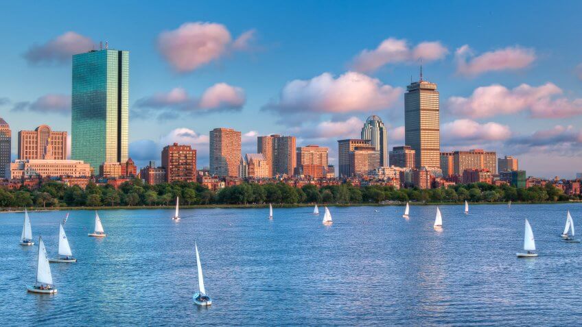 A panoramic view of the full Boston skyline with the Charles River full of sailboats as sunset approaches.