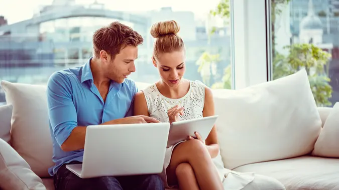 Young couple sitting on sofa in an apartment and using digital tablet and laptop together.