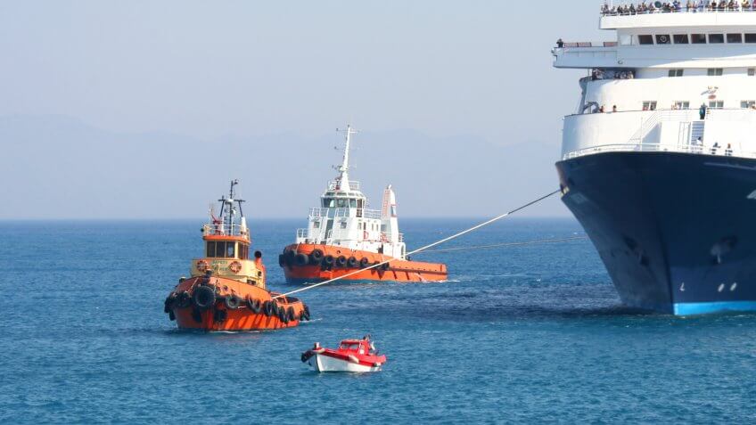 Tugboats and a pilot boat maneuvering a large tourist ferry into a harbour on the Greek island of Rhodes.