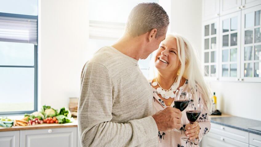 Affectionate senior couple drinking wine in the kitchen together with focus to the smiling attractive wife looking lovingly at her husband.