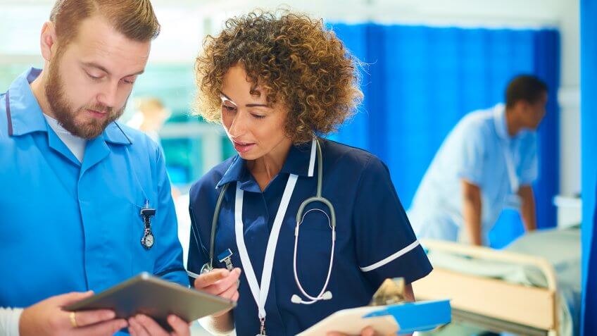 a nursing sister stands with a male nurse chatting about the medical chart that she is holding and cross referencing it with the digital tablet that he is holding.