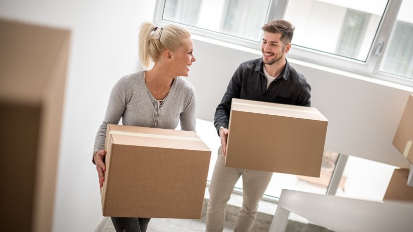 Couple carrying boxes upstairs through their new building and smiling.