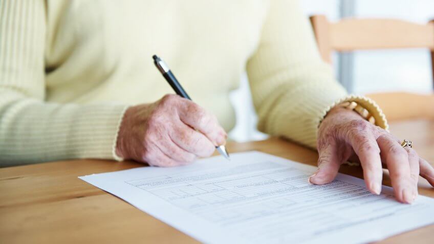 A cropped shot of an elderly woman sitting at a table and signing a document.