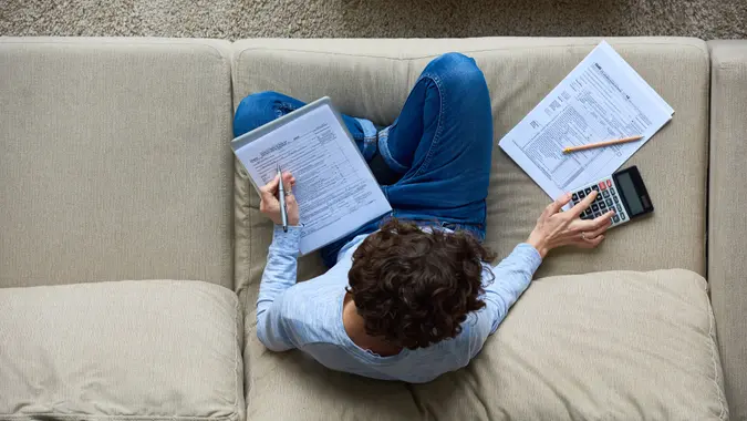 Directly above view of concentrated accountant using calculator while preparing tax forms sitting on sofa.