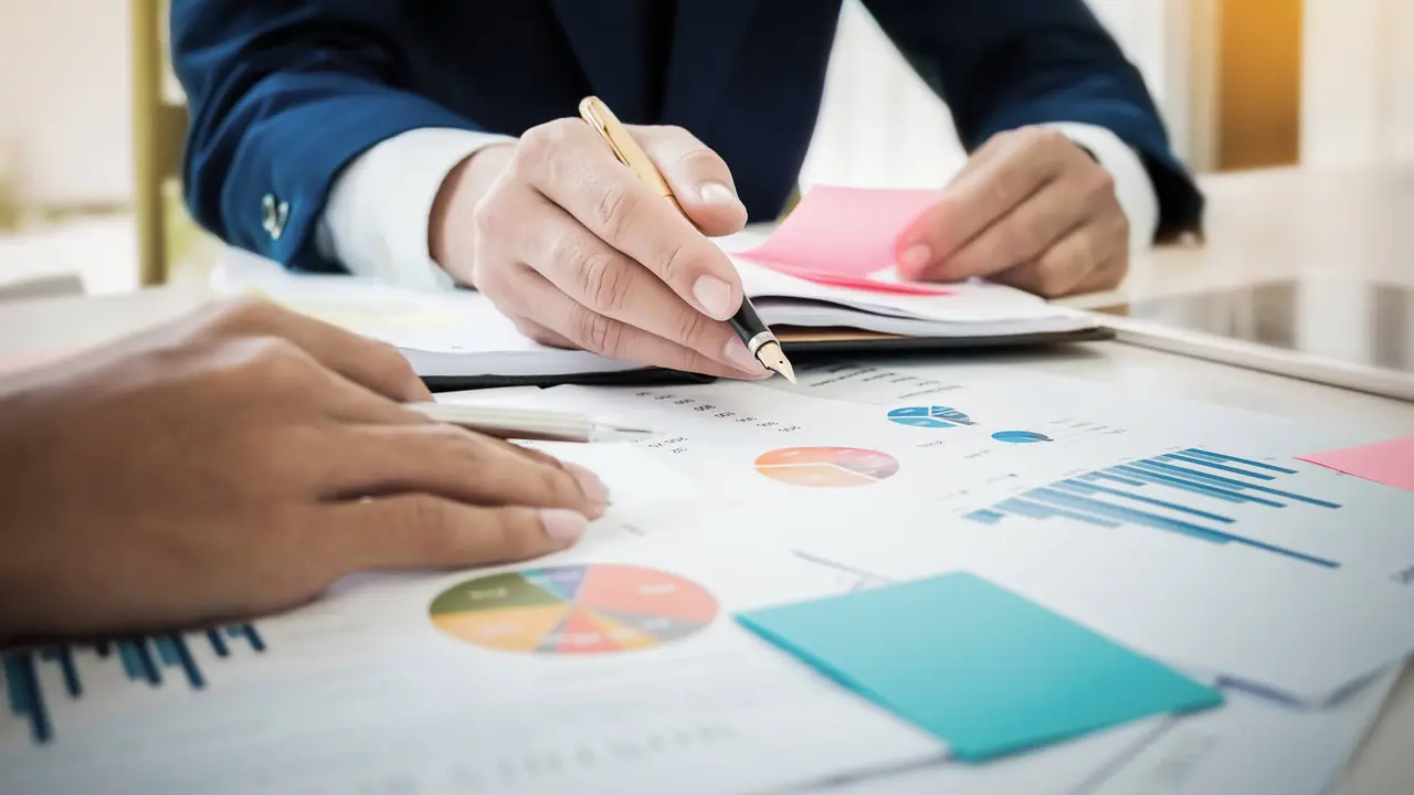 Young business man hand holding pen pointing graph data documents and partner working at office with tablet,post it note, for Plans to improve quality next month on his desk.